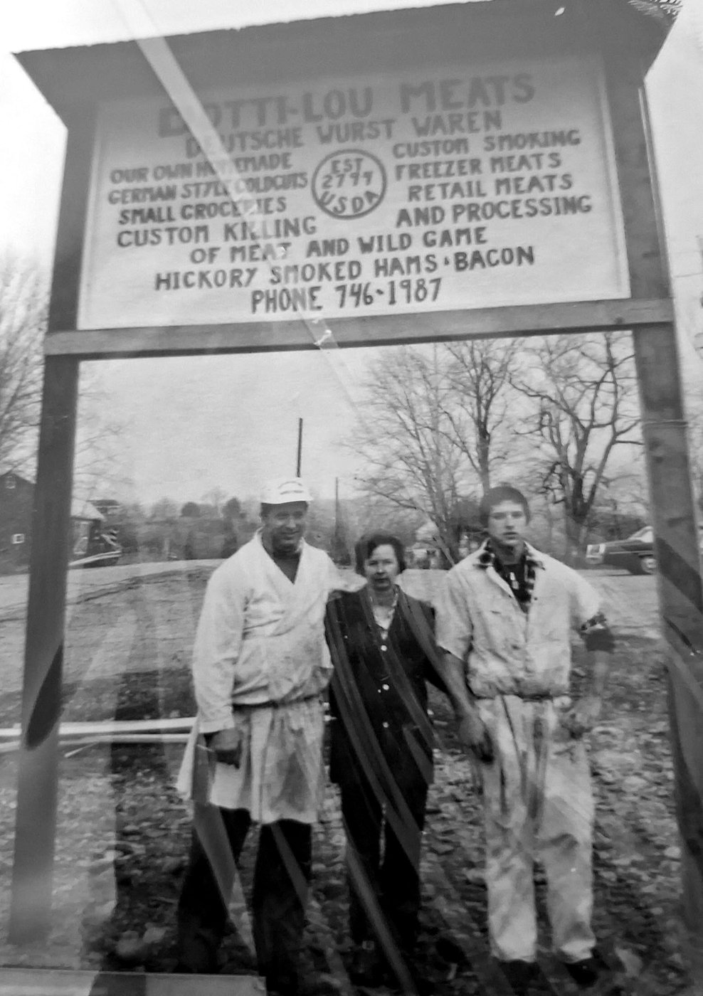 An early photograph of three people standing beneath the hand-lettered Dotti-Lou Meats sign.