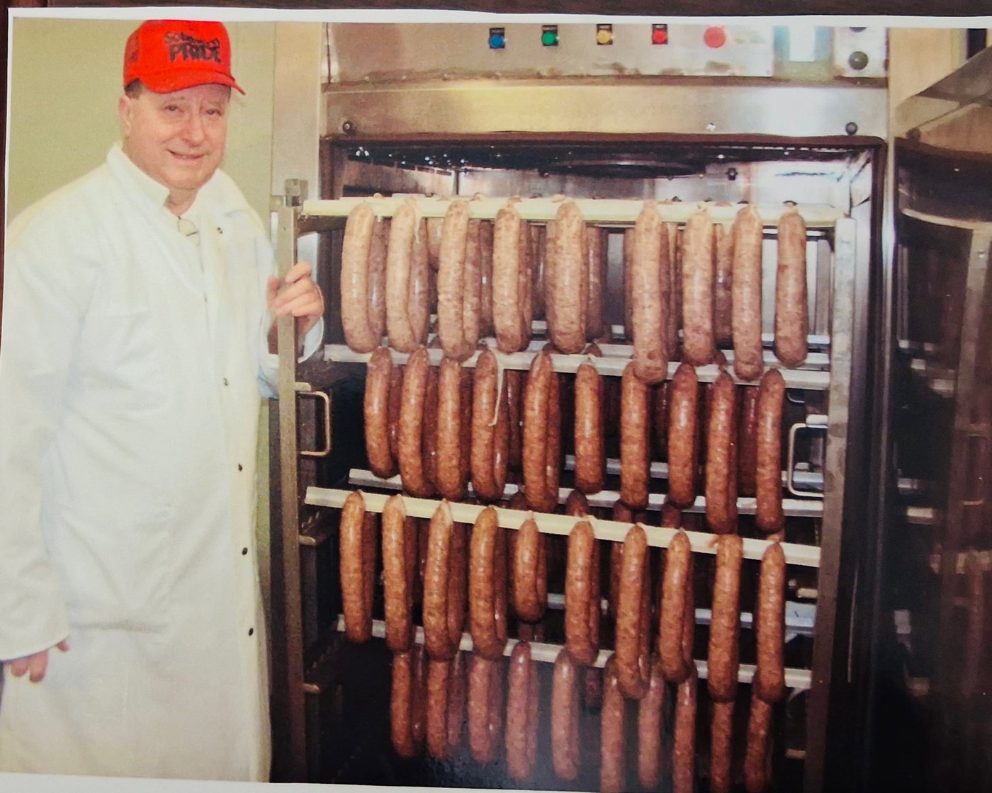 A butcher in a white coat standing beside racks of sausage hanging in a smokehouse.