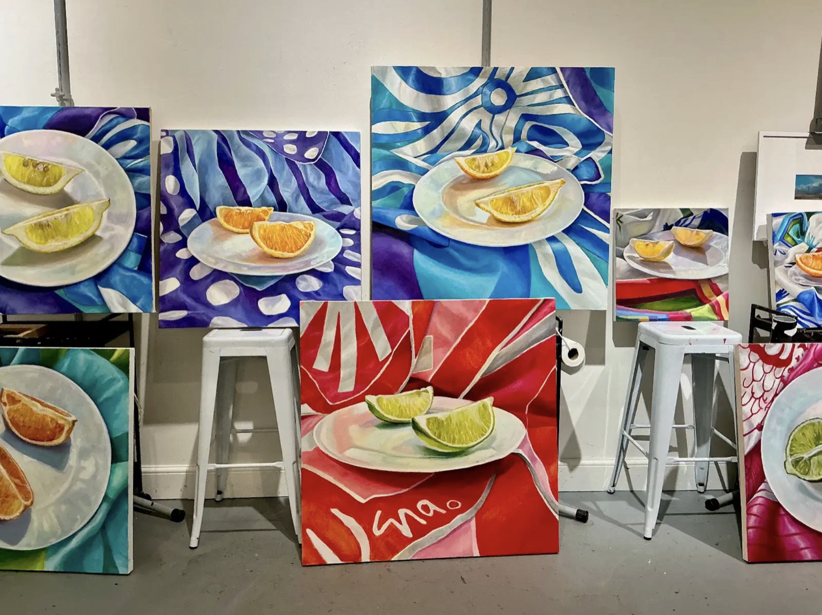 Nine finished square oil paintings leaning against a studio wall, each showing citrus wedges on a white plate over a brightly patterned scarf.