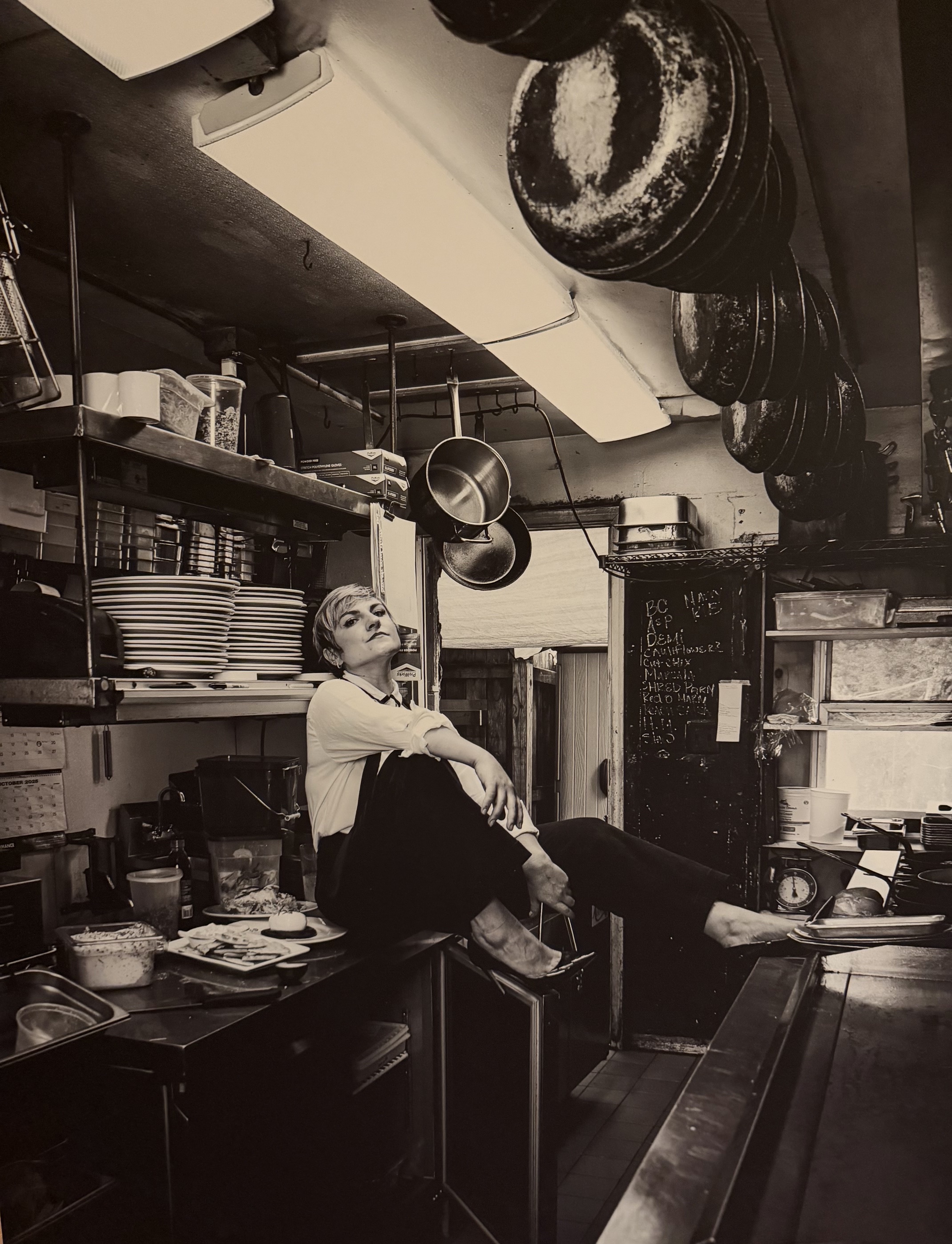 Emily Fischer seated on a stainless steel counter in the Limeport Inn kitchen, surrounded by stacked plates and hanging pans.