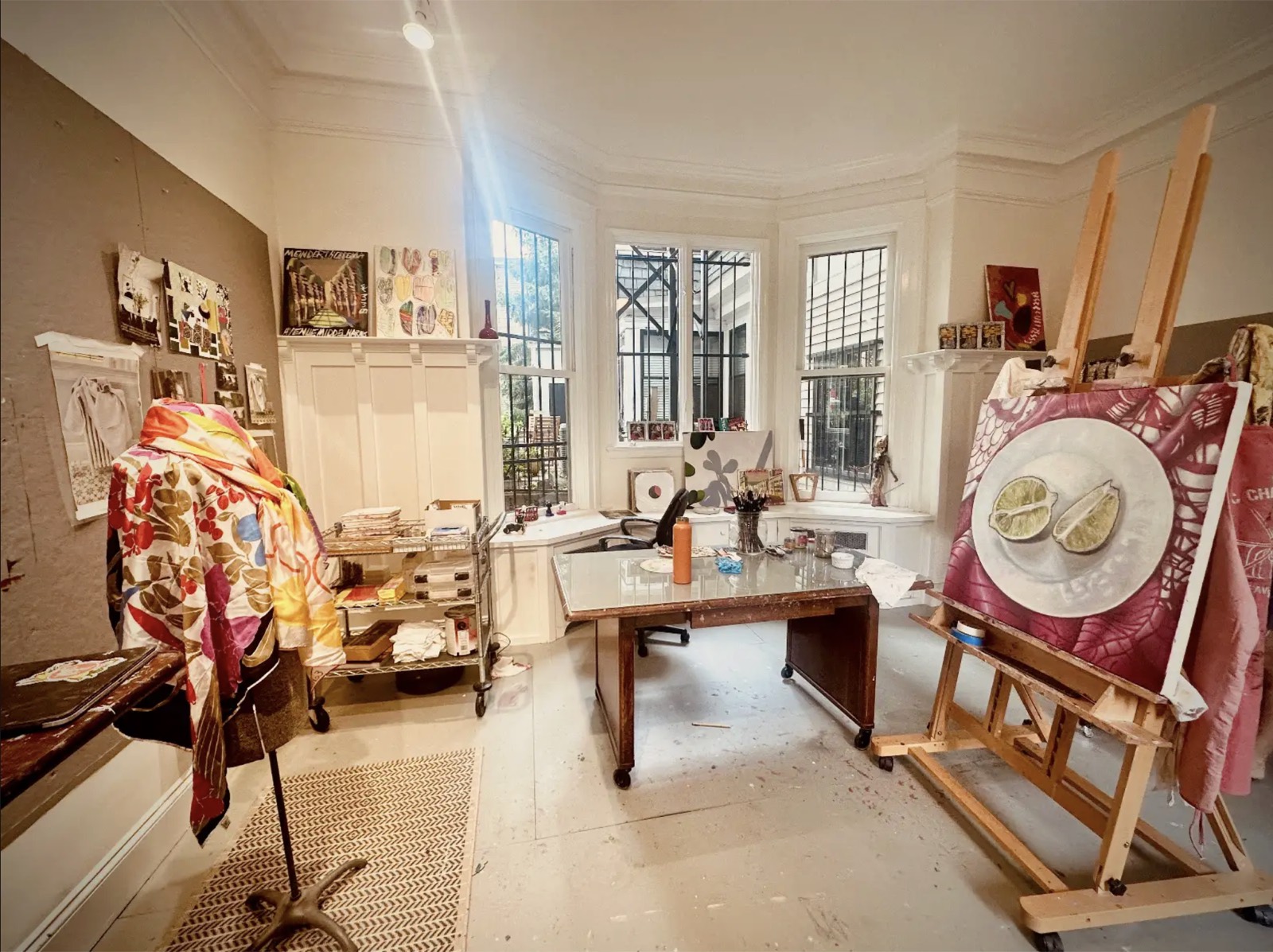 A bright studio with bay windows, a scarf draped over a stand for reference, a worktable of brushes, and a lime-on-a-plate painting in progress on a wooden easel.