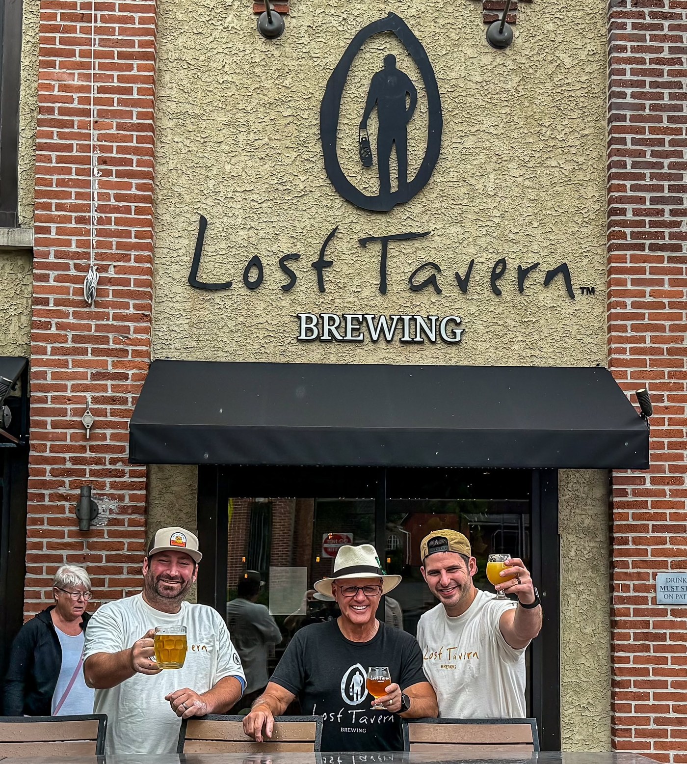 The Lost Tavern Brewing team raising glasses outside the Hellertown taproom beneath the Lost Tavern Brewing sign