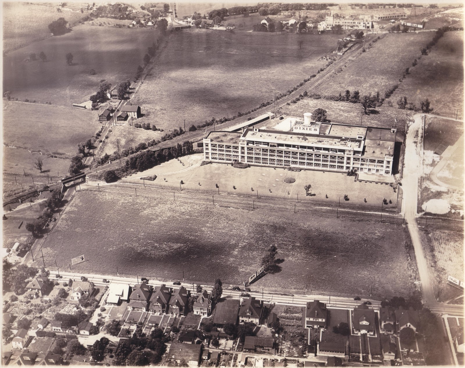 Early aerial view of the Dixie Cup plant standing alone among farm fields, with the rail line and rows of houses below