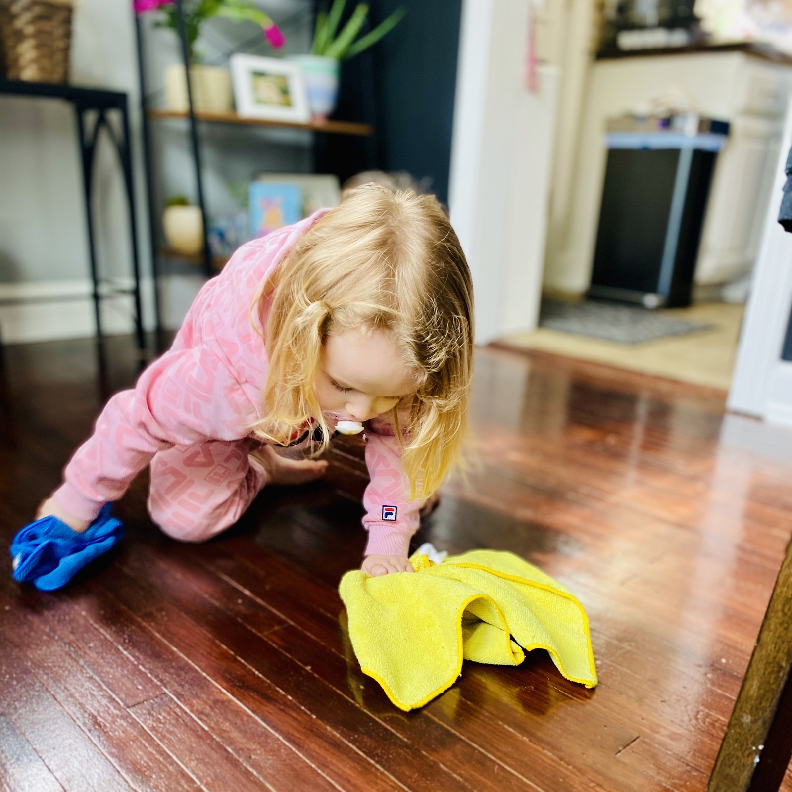 A small child in pink pyjamas wiping a hardwood floor with a yellow cloth.