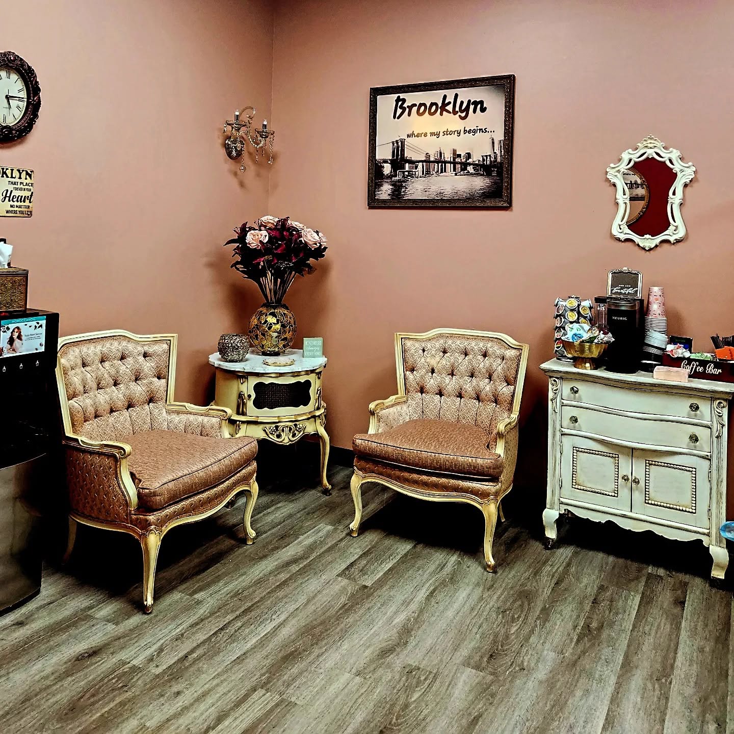 The salon waiting area: two tufted armchairs in gilt frames, an antique side table with flowers, a coffee bar on a white dresser, and a framed Brooklyn Bridge print on a dusty pink wall.