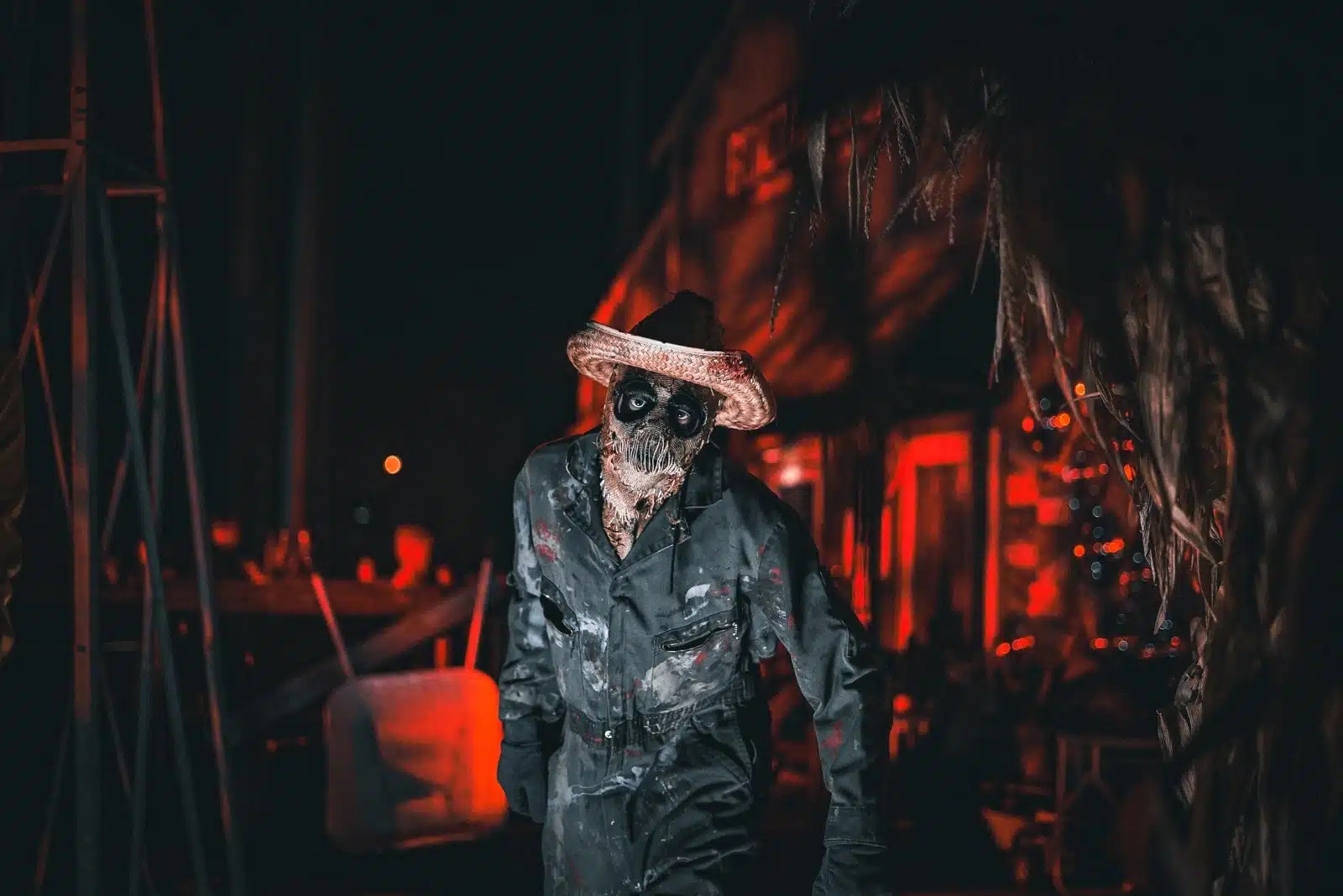 A costumed scareactor in a straw hat and work coveralls standing in a dark, red-lit scare zone