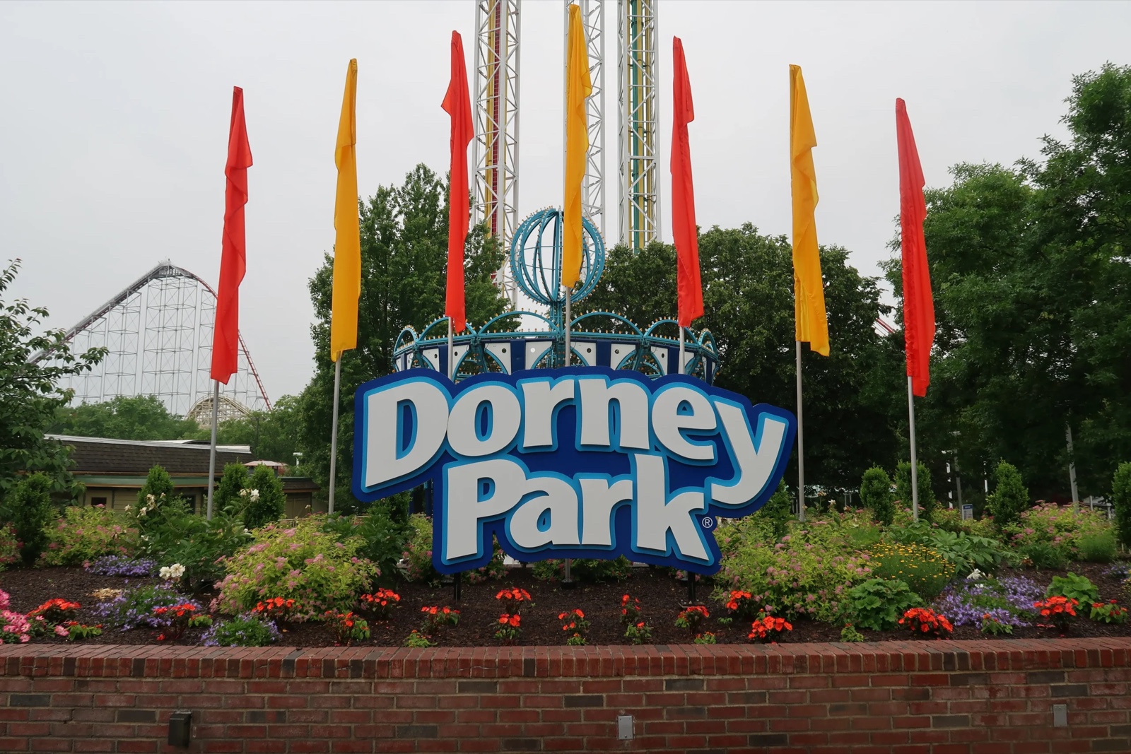 The Dorney Park entrance sign in daylight, with flags, flower beds and a roller coaster behind it