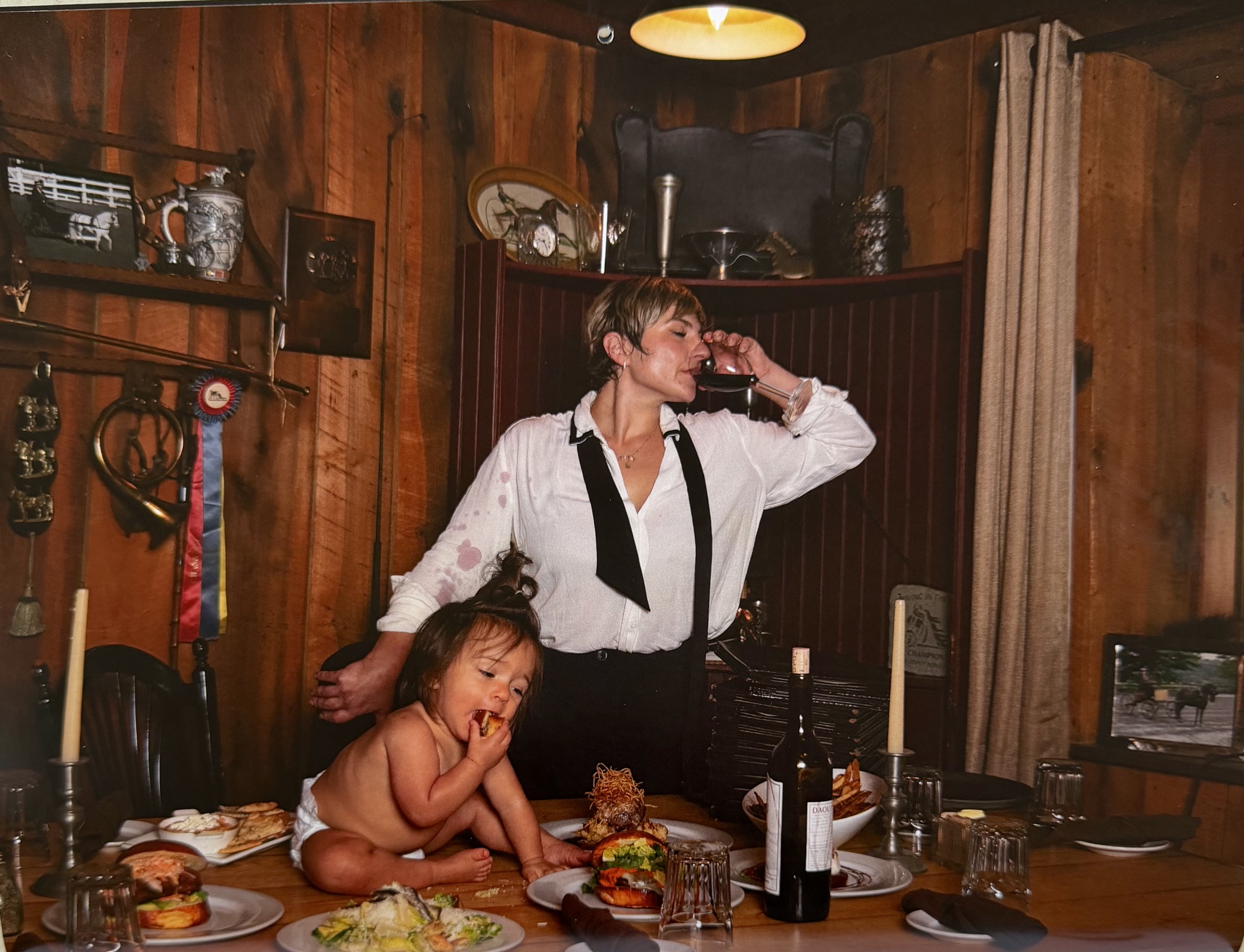 Emily Fischer standing at a set dining table with a glass of wine while her daughter Iris sits on the table eating a piece of bread.