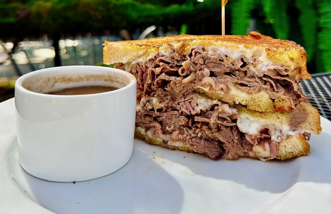 A sandwich of shaved beef and melted cheese on grilled bread, cut in half, with a cup of jus alongside.