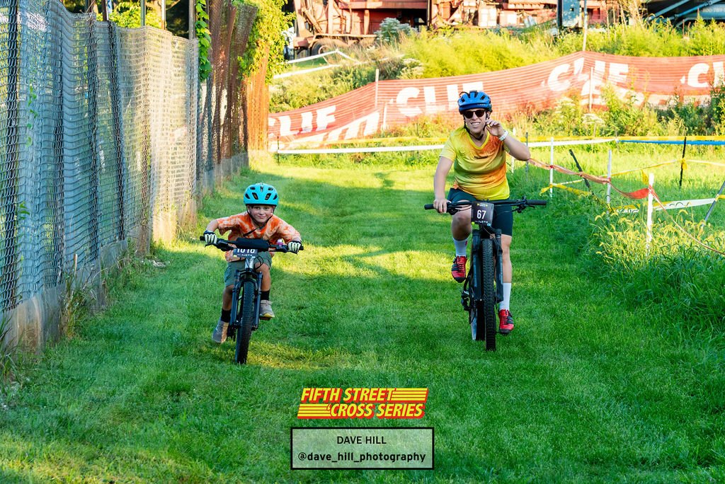 A smiling boy and an adult ride bikes side by side along a grassy course