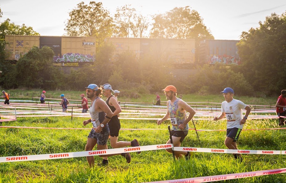 Runners cross a grassy field at sunset as a freight train passes behind them
