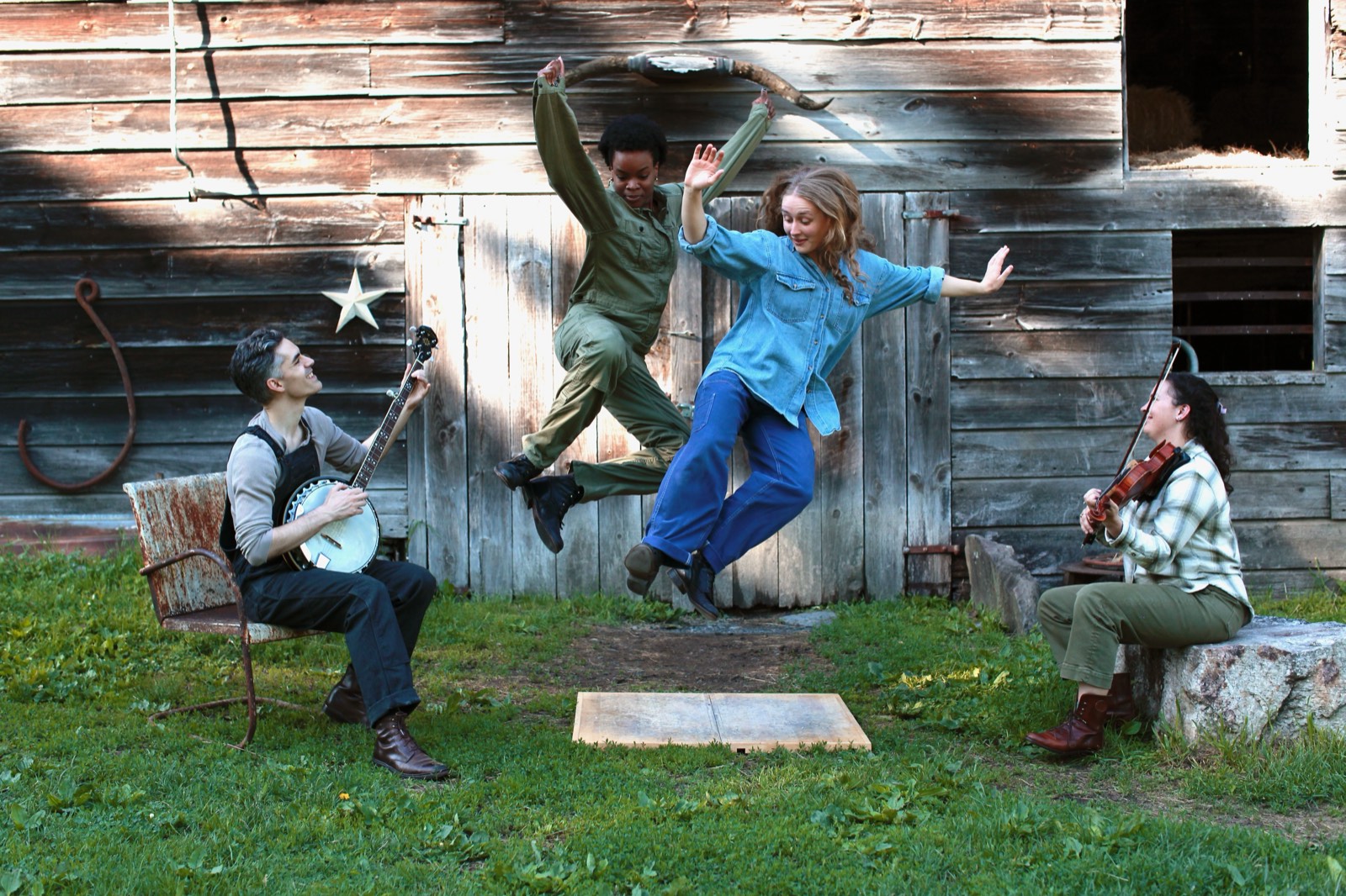 Three Vanaver Caravan dancers in work clothes lean back in unison inside a weathered barn, light coming through the gaps in the boards
