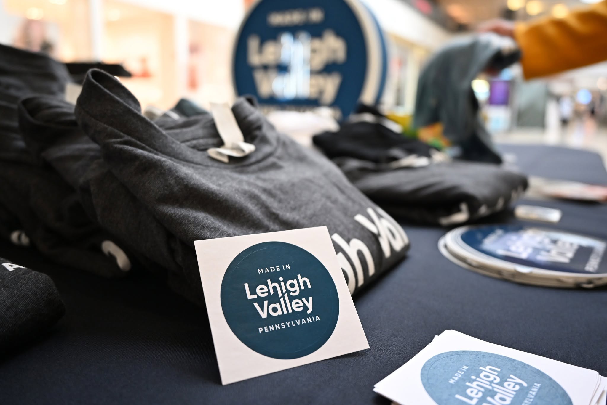 A Made in Lehigh Valley Pennsylvania sticker propped against folded branded t-shirts on a giveaway table, with the new circular sculpture out of focus behind it.