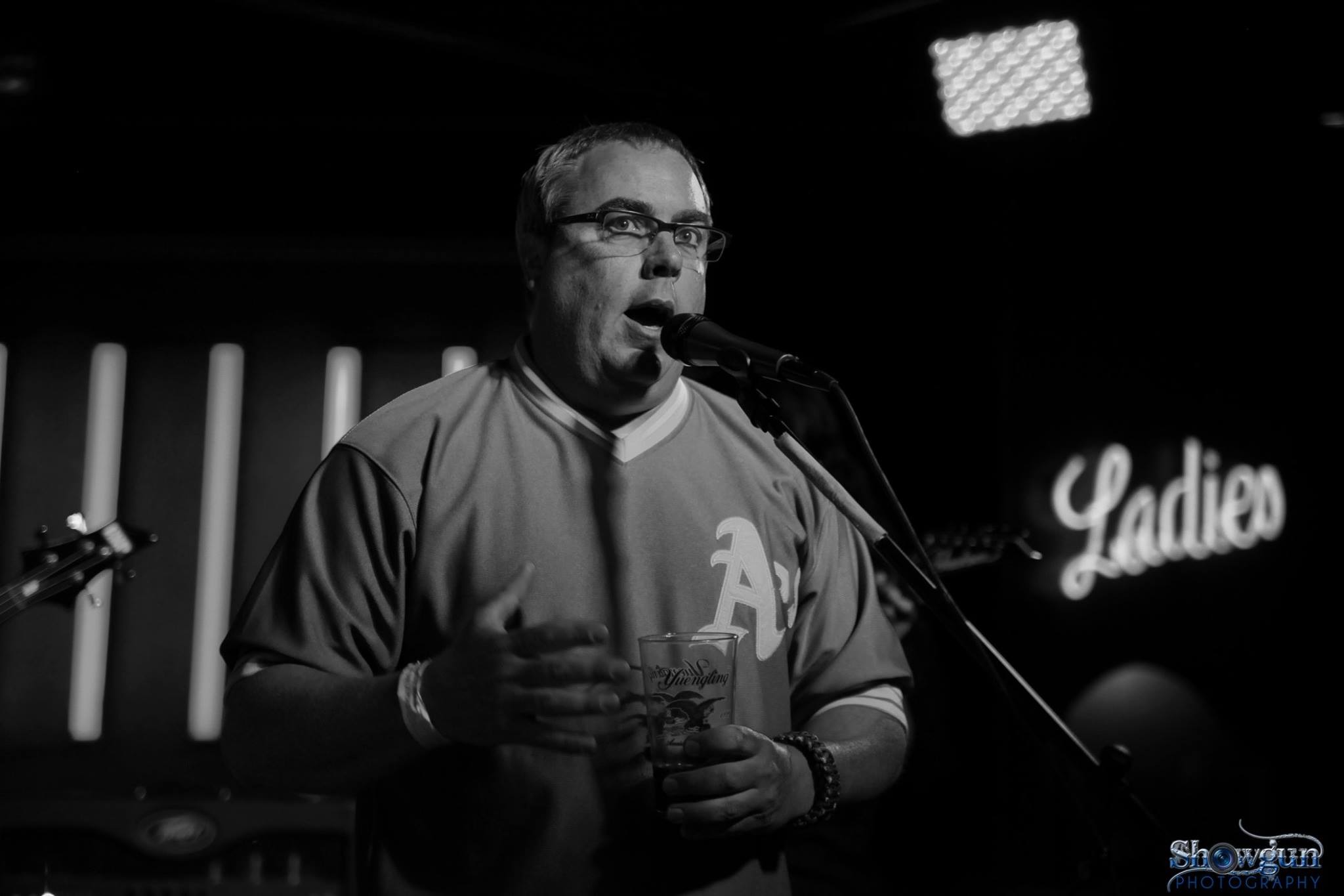 Matt Metal speaking into a microphone on a club stage, holding a pint glass, with a guitar and amplifier behind him