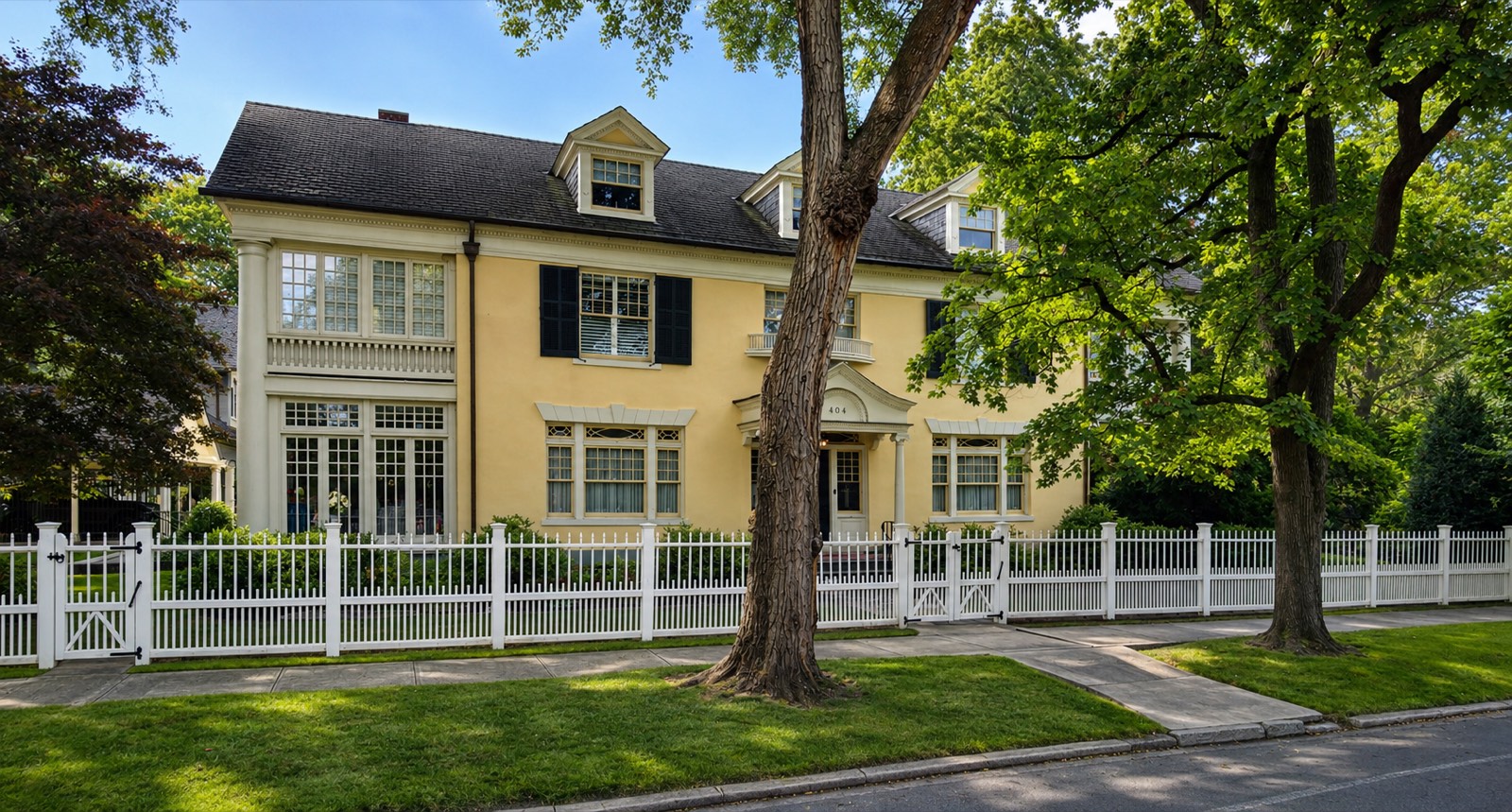 A yellow stucco Colonial Revival house with black shutters, dormers and a white picket fence